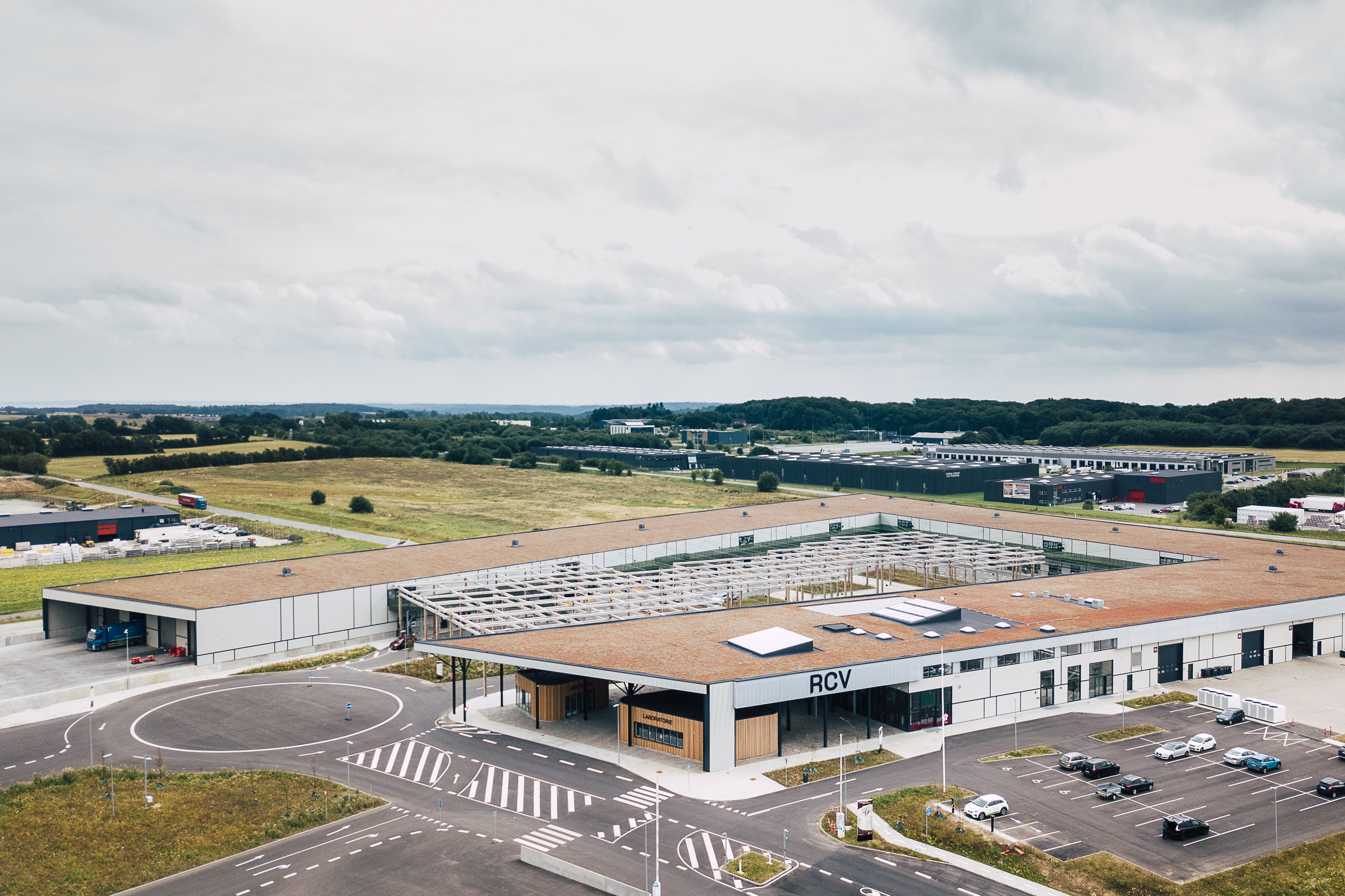 Luftfoto af RessourceCenter Vejle (RCV). Centret har en stor bygning med fladt tag, en parkeringsplads og tydeligt markerede vejbaner og indkørsler. Taget har solpaneler, og der er en åben konstruktion med trærammer i gårdområdet.